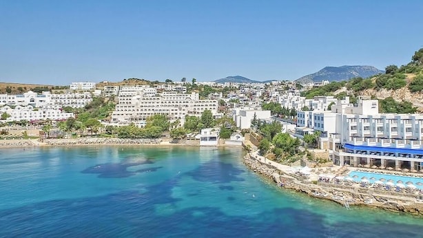 Un pueblo costero con edificios a lo largo del paseo marítimo, un mar azul cristalino y una montaña al fondo.