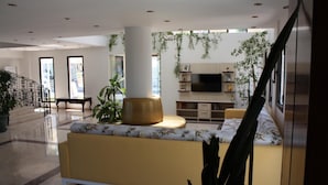 Un hall espacioso con un sofá amarillo, una planta grande y un televisor montado en la pared.