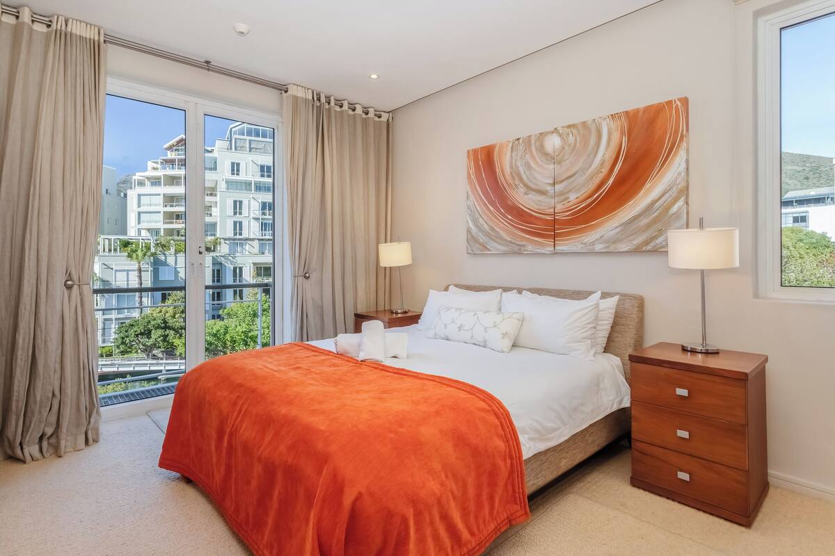 Ein Schlafzimmer mit einem großen Bett, zwei Nachttischen, einem Beistelltisch, einem Fenster mit Vorhängen und Blick auf die Stadt.