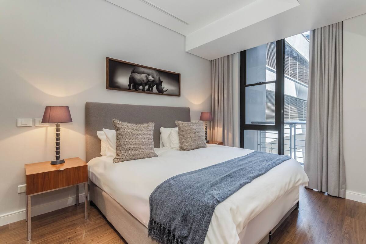 Une chambre moderne avec un grand lit, deux tables de chevet avec des lampes, une couverture grise et un tableau encadré représentant des rhinocéros accroché au mur.