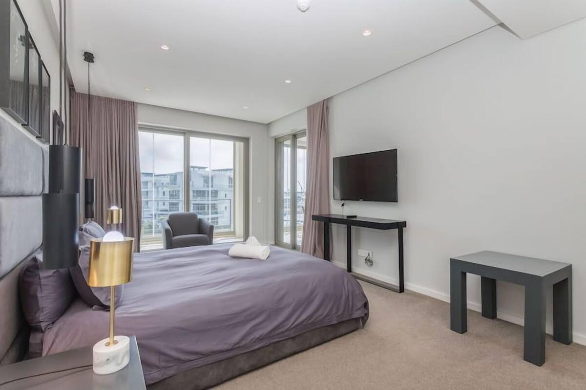 Une chambre d’hôtel moderne dotée d’un grand lit, d’un téléviseur à écran plat, d’une table de chevet et offrant une vue sur la ville grâce à de grandes fenêtres.
