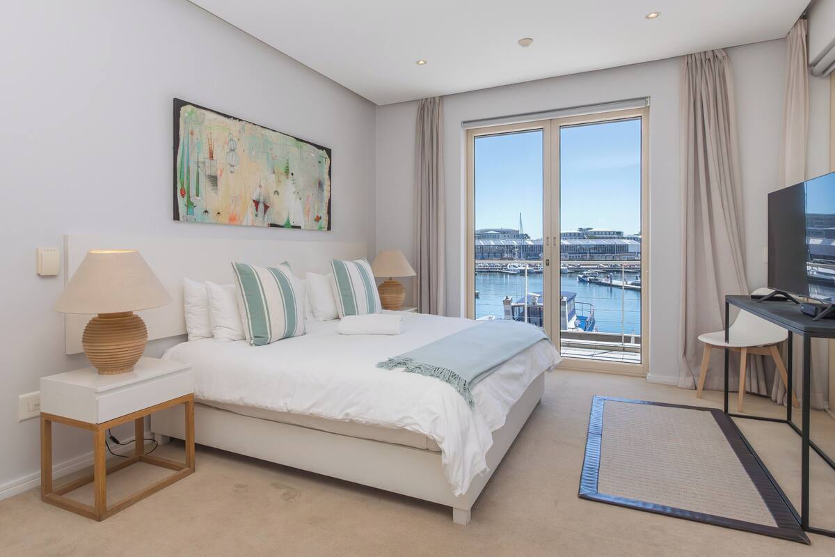 Ein modernes Hotelzimmer mit einem großen Bett, einem Schreibtisch, einem Fernseher und Blick auf den Hafen.