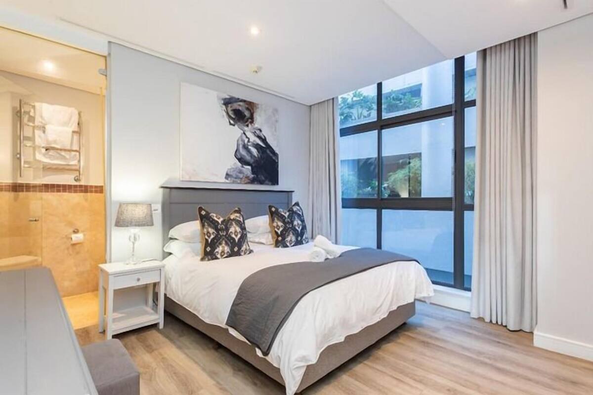 Une chambre d’hôtel moderne avec un grand lit, une table de chevet, une commode, une fenêtre avec des rideaux et un espace douche.