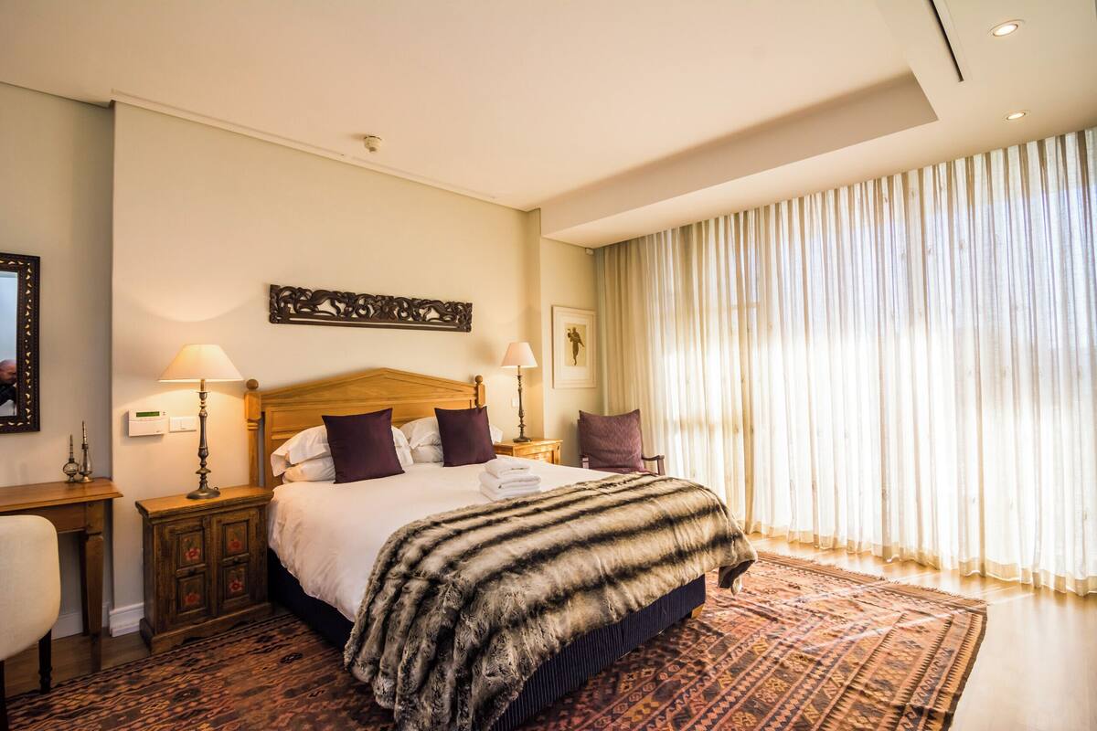 Une chambre d’hôtel avec un grand lit, deux tables de chevet avec des lampes, une tête de lit, un tapis à motifs et une fenêtre avec des rideaux.