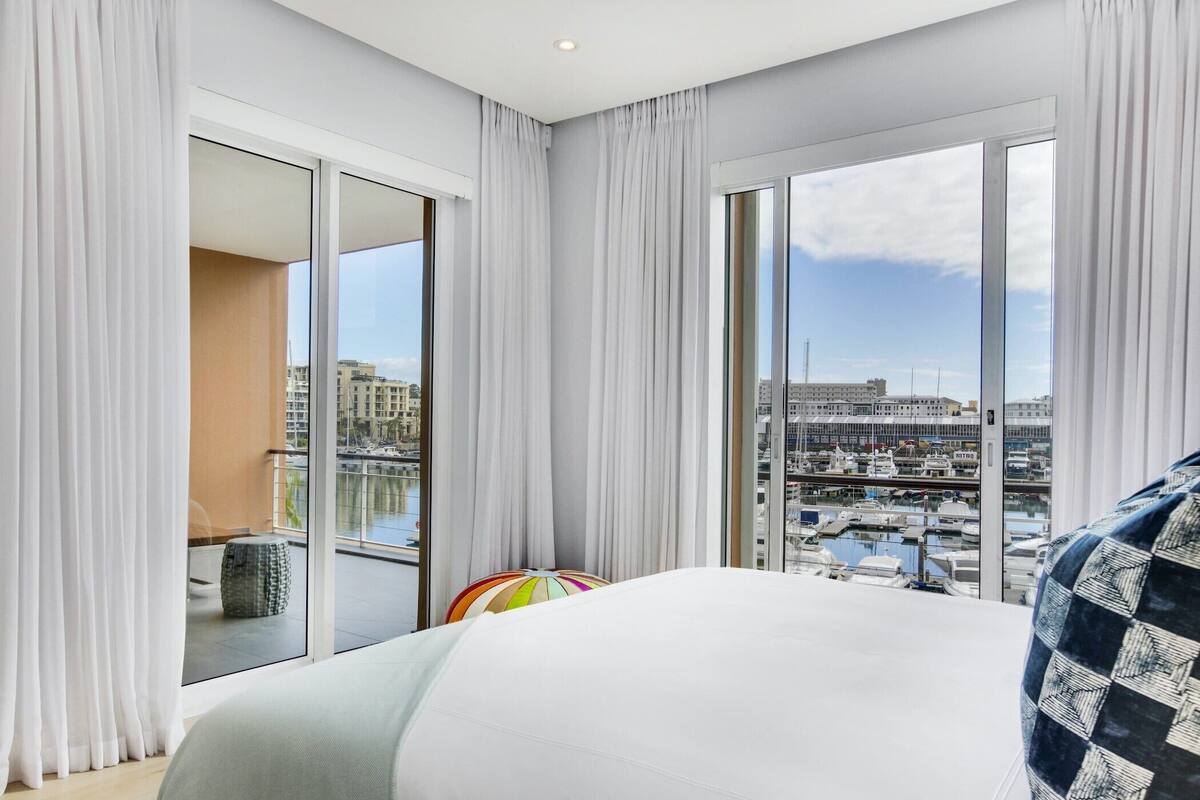 Une chambre d’hôtel moderne avec un grand lit, un oreiller coloré et une vue sur un port de plaisance et des immeubles, grâce à de grandes fenêtres du sol au plafond.