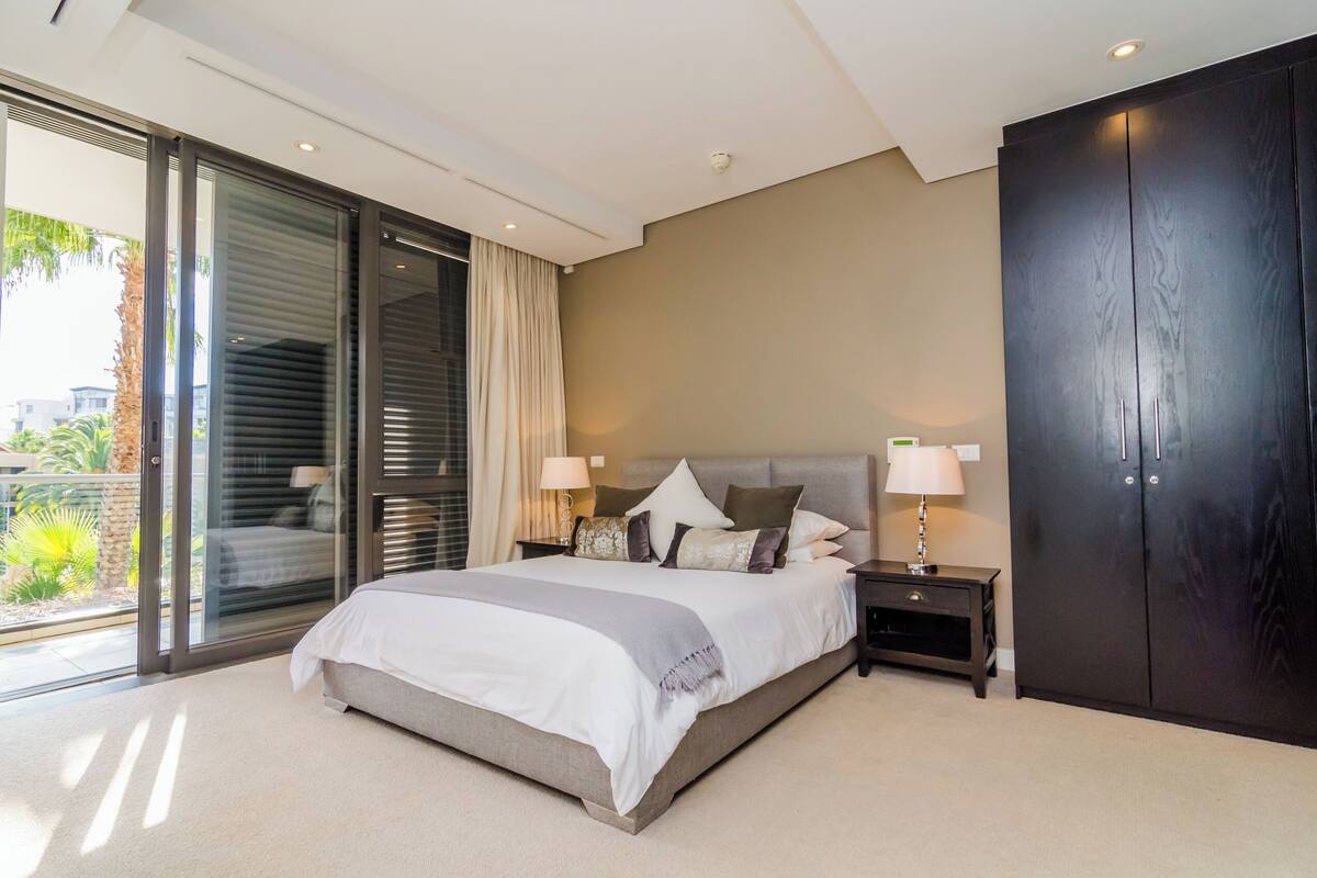 Une chambre moderne avec un grand lit, une table de chevet, une armoire sombre et une porte-fenêtre coulissante donnant sur un balcon.