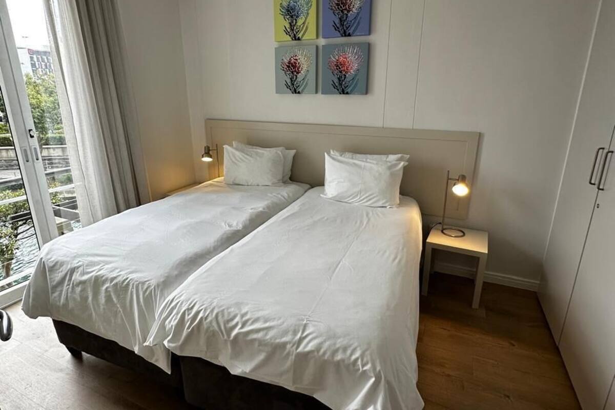 Ein Doppelbett mit weißer Bettwäsche, zwei Nachttische mit Lampen und Blick auf Grün hinter einem Fenster.