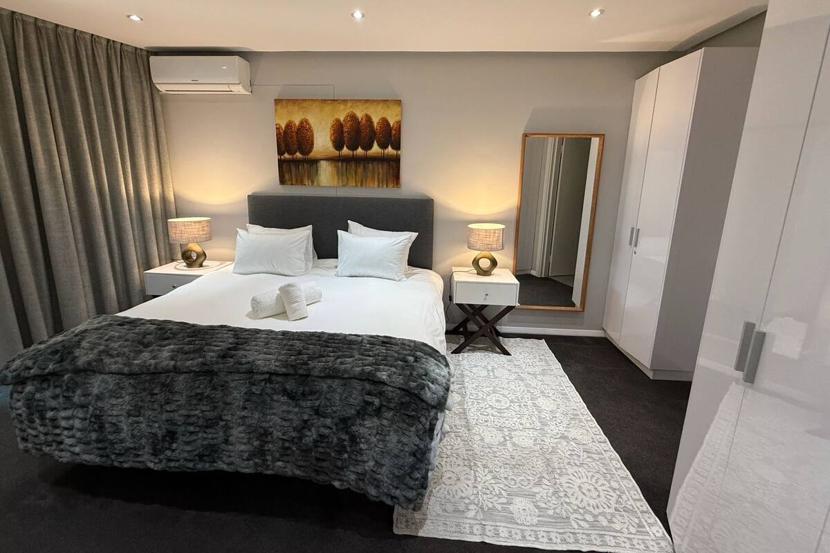 Une chambre d’hôtel moderne avec un grand lit, deux tables de chevet avec des lampes, un grand miroir et une armoire.
