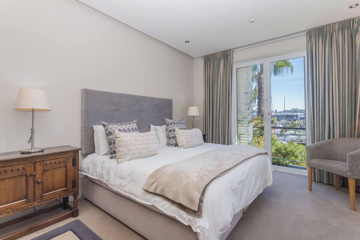 Une chambre moderne avec un grand lit, une table de chevet en bois, une chaise et une vue sur la ville grâce à de grandes fenêtres.