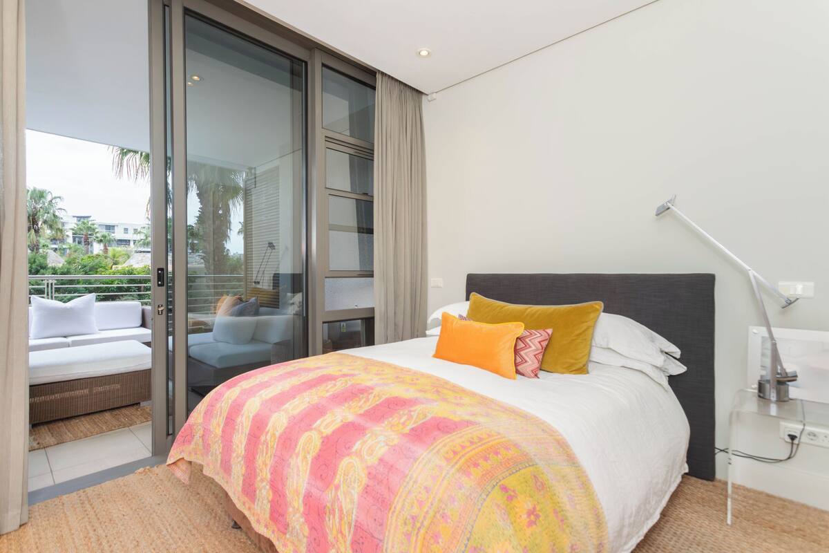 Une chambre dotée d’un grand lit, d’une table de chevet, d’une lampe et d’une porte-fenêtre coulissante donnant sur un balcon aménagé avec des sièges d’extérieur.
