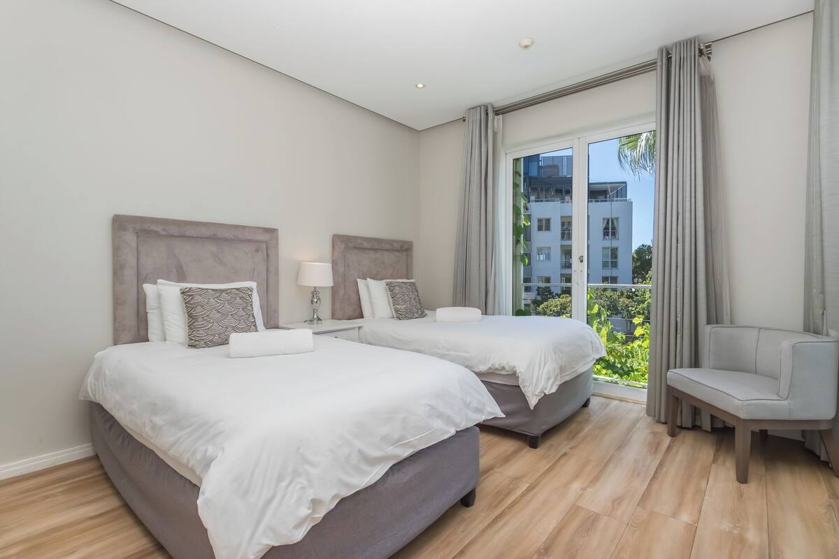 Une chambre d’hôtel avec deux lits, une chaise, une fenêtre avec des rideaux et une vue sur des bâtiments et de la verdure.