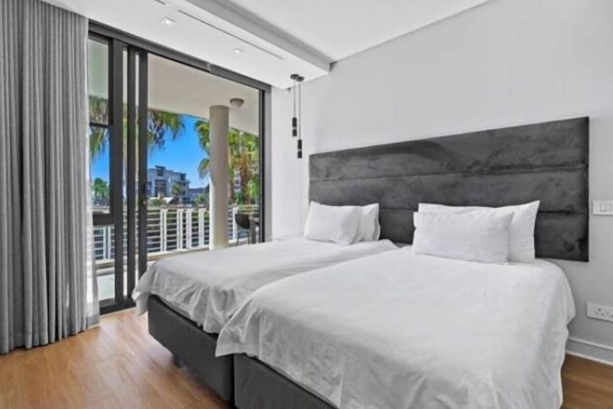Une chambre d’hôtel moderne avec deux lits, une grande porte-fenêtre coulissante donnant sur un balcon, et une vue sur des palmiers et des immeubles.