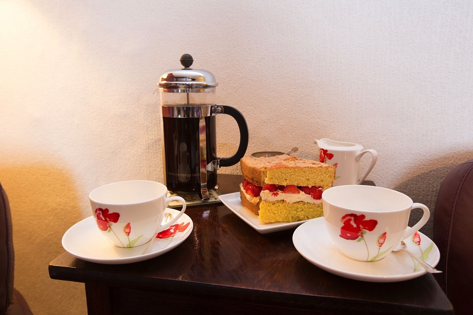 A table with a coffee maker, two cups with floral designs, and a slice of cake with strawberries.