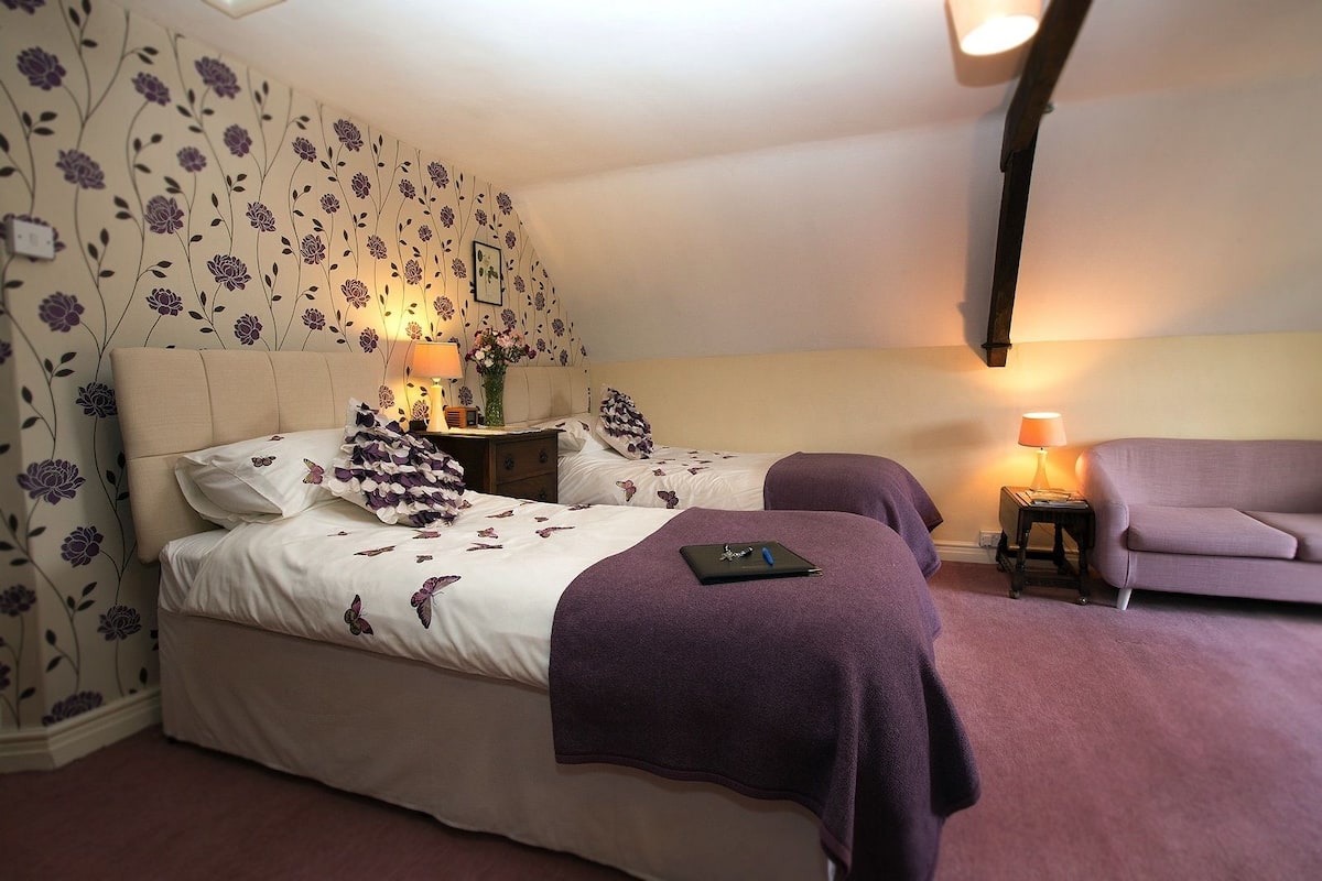 A bedroom with two beds, a nightstand, a chair, and a wall with floral wallpaper.