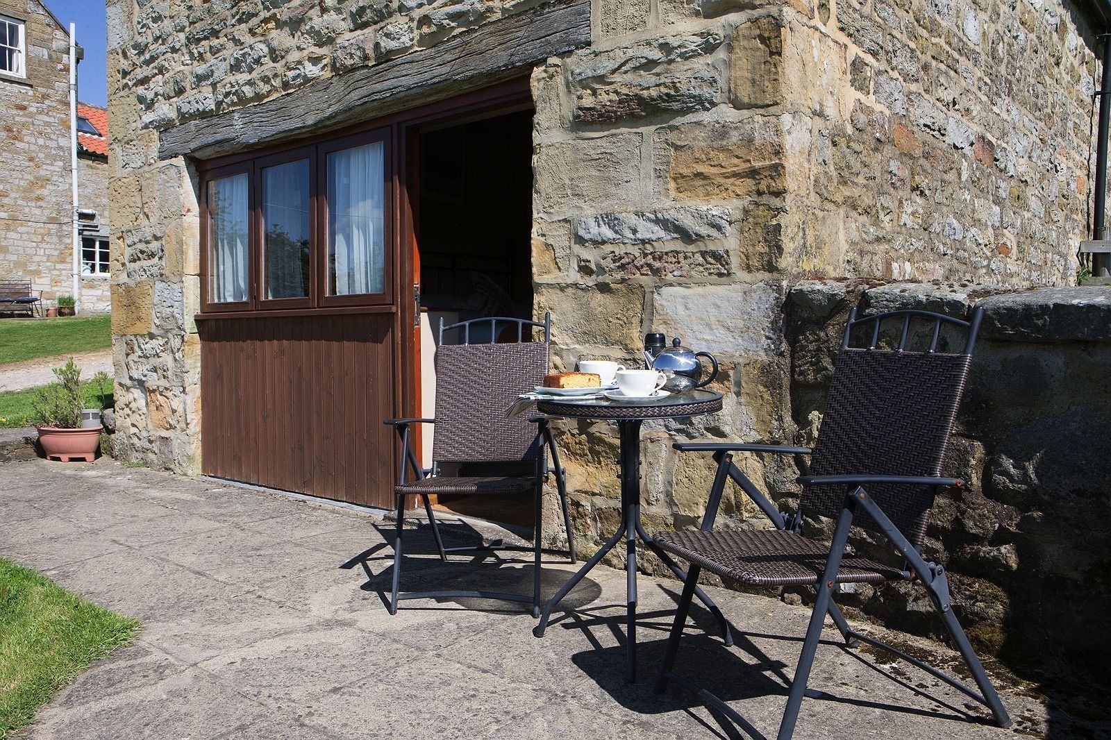 A stone building with a wooden door and windows, a small outdoor table with chairs, and a potted plant.