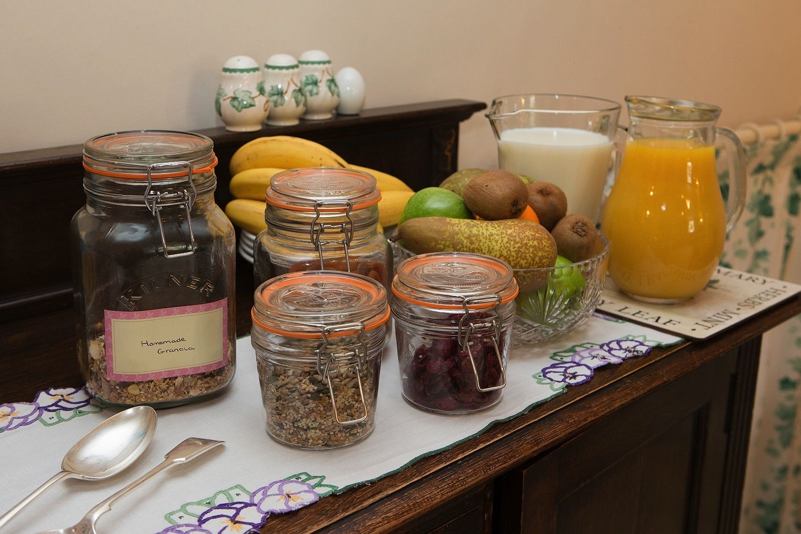 Una encimera de cocina con frascos de granola casera, frutas frescas y una jarra de jugo de naranja.