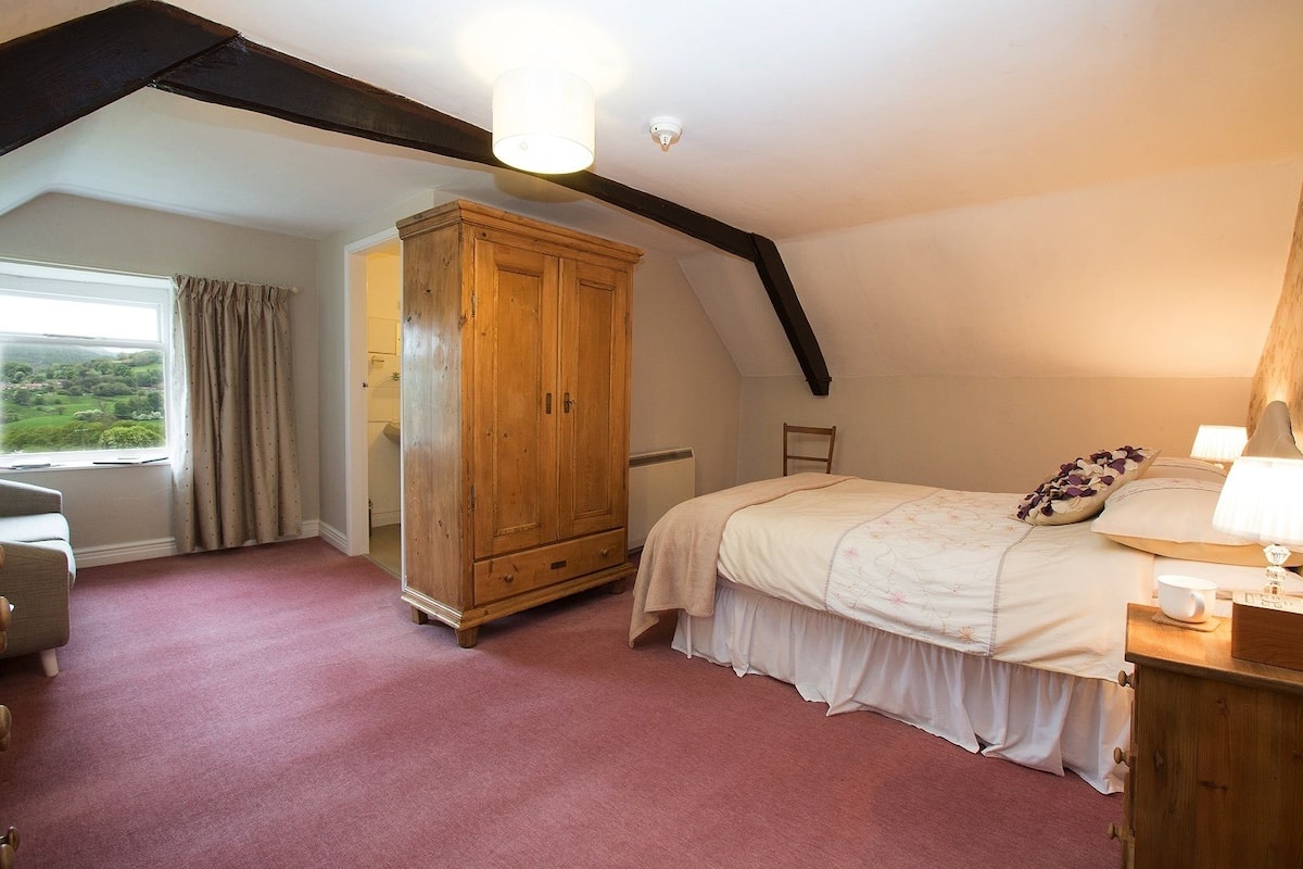 A bedroom with a large wooden wardrobe, a bed with a white duvet, a nightstand with a lamp, and a window with curtains.