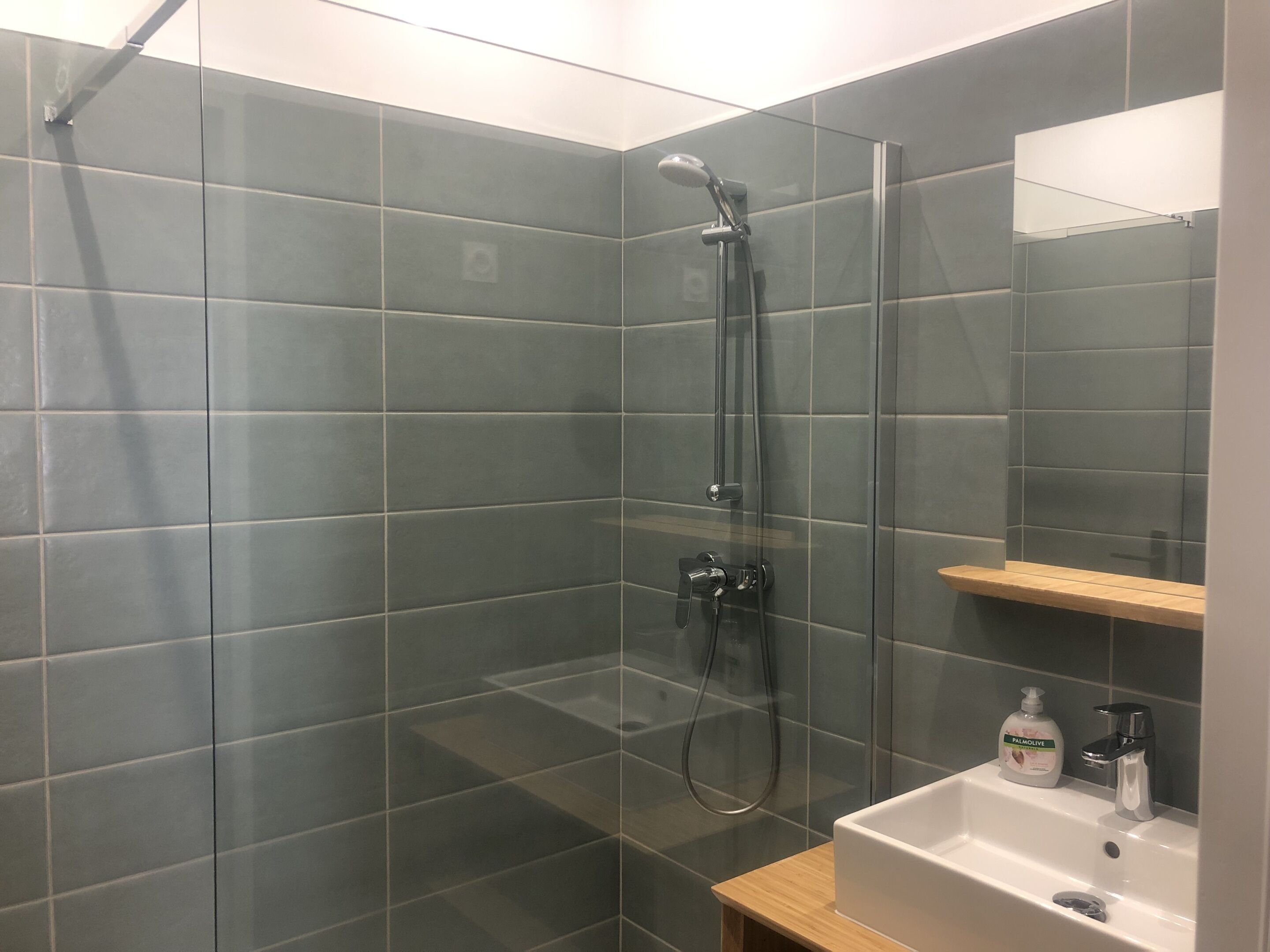 Un baño moderno con ducha envuelta en vidrio, un lavabo blanco y un estante de madera.