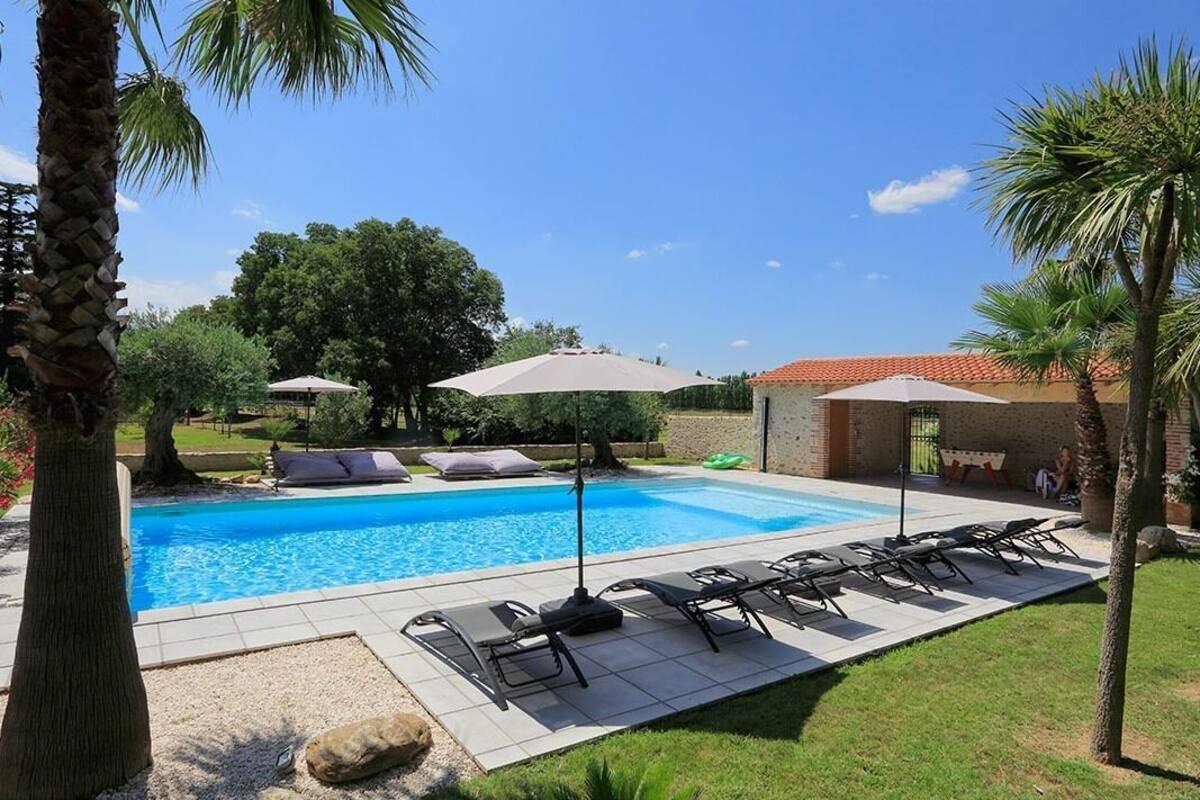 Ein Swimmingpool mit Liegestühlen und Sonnenschirmen, umgeben von Palmen und einem Backsteinhaus.