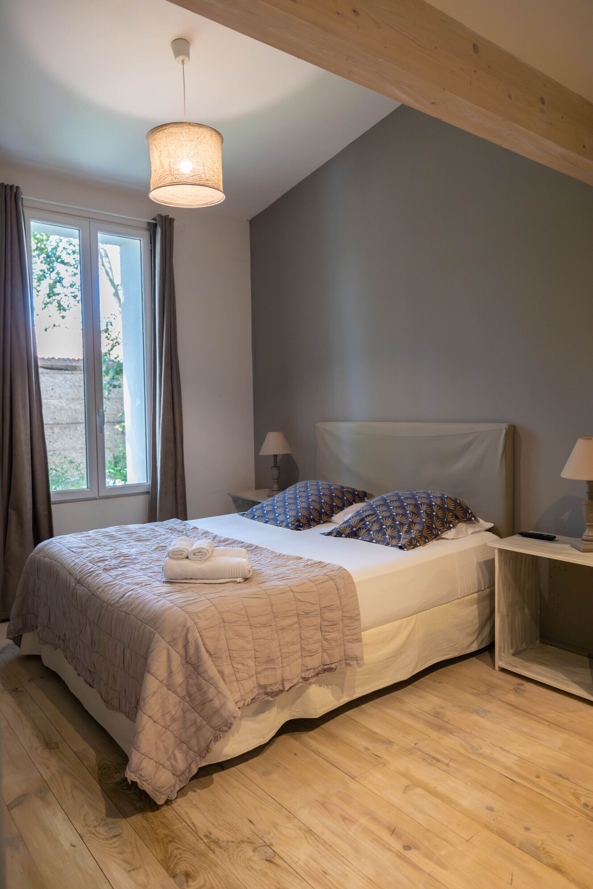 Quarto com cama, mesa de cabeceira, luminária e janela com cortinas.