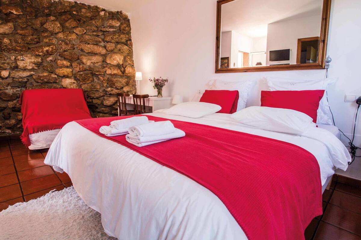 Une chambre d’hôtel avec un mur en pierre, un lit avec une literie blanche et rouge, une chaise rouge, une petite table avec une lampe et un grand miroir.