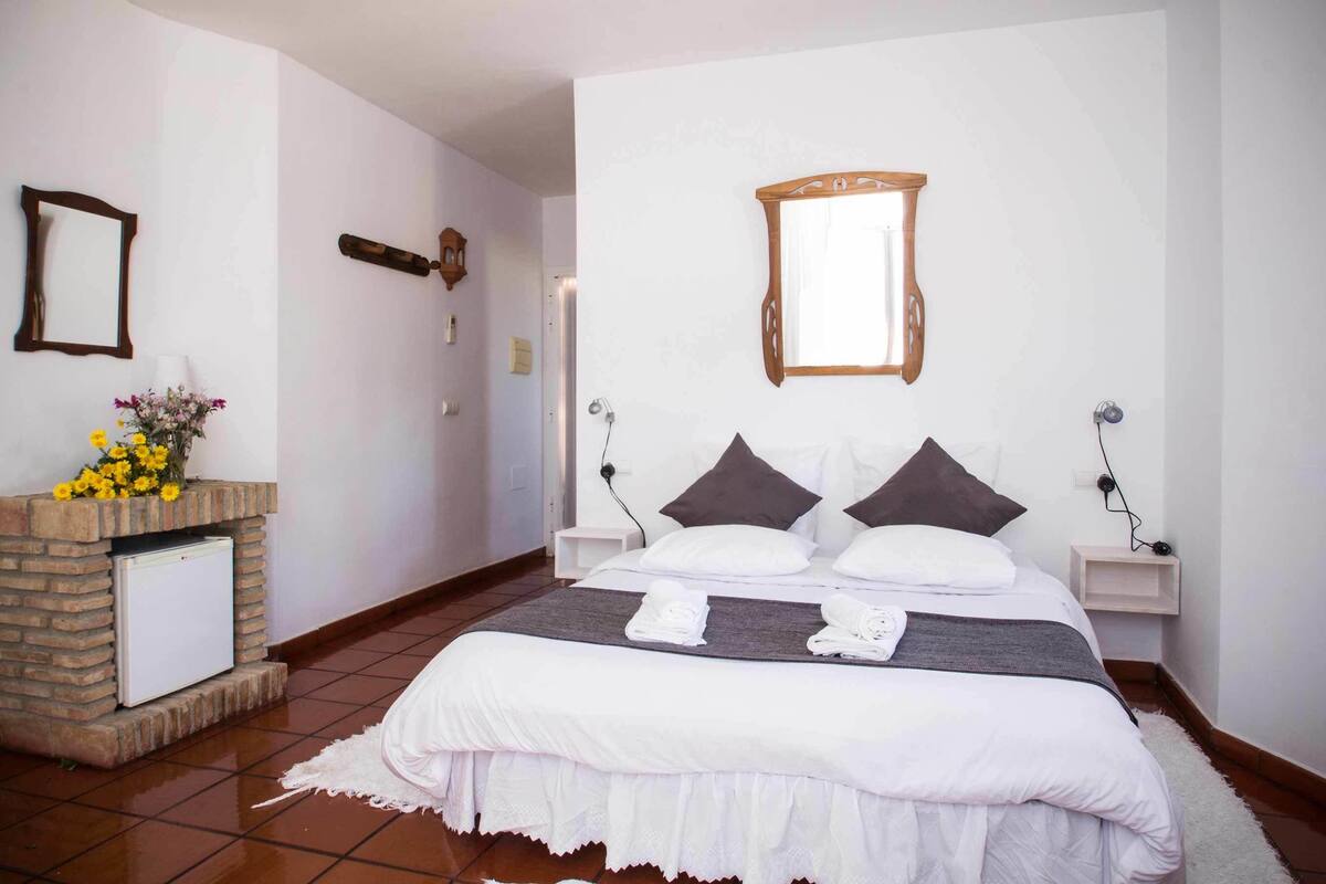 Un grand lit recouvert d’une courtepointe blanche et agrémenté de deux oreillers, une petite table de chevet avec une lampe, un miroir fixé au mur et une cheminée en briques surmontée d’un vase de fleurs.