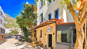 A street view with a bakery named "Baden" on the right, a tree with hanging bread, and a mountain in the background. - Bahar Pansiyon (Kas)