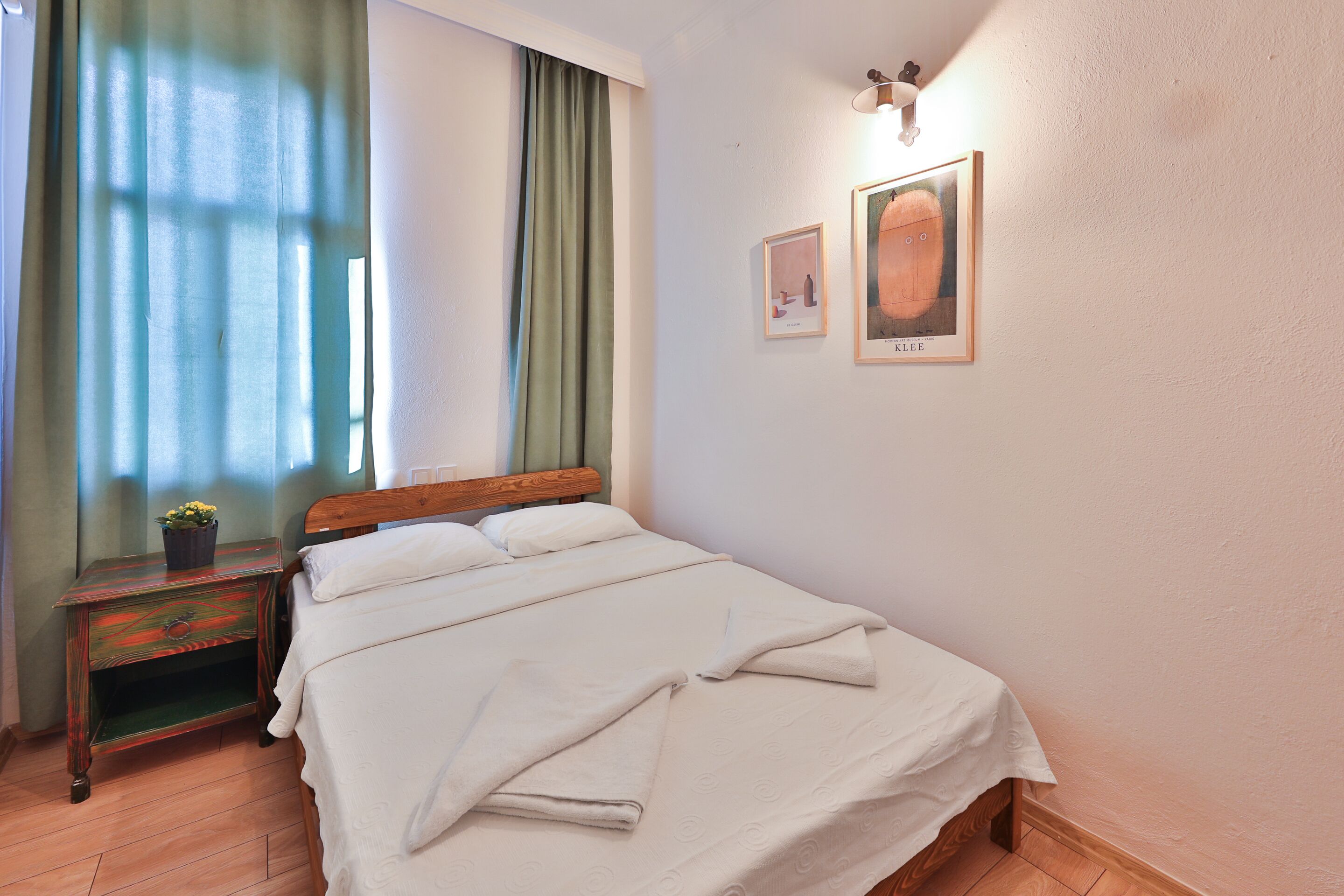 A neatly made bed with a white bedspread and pillows, a wooden nightstand with a plant, and two framed pictures on the wall.