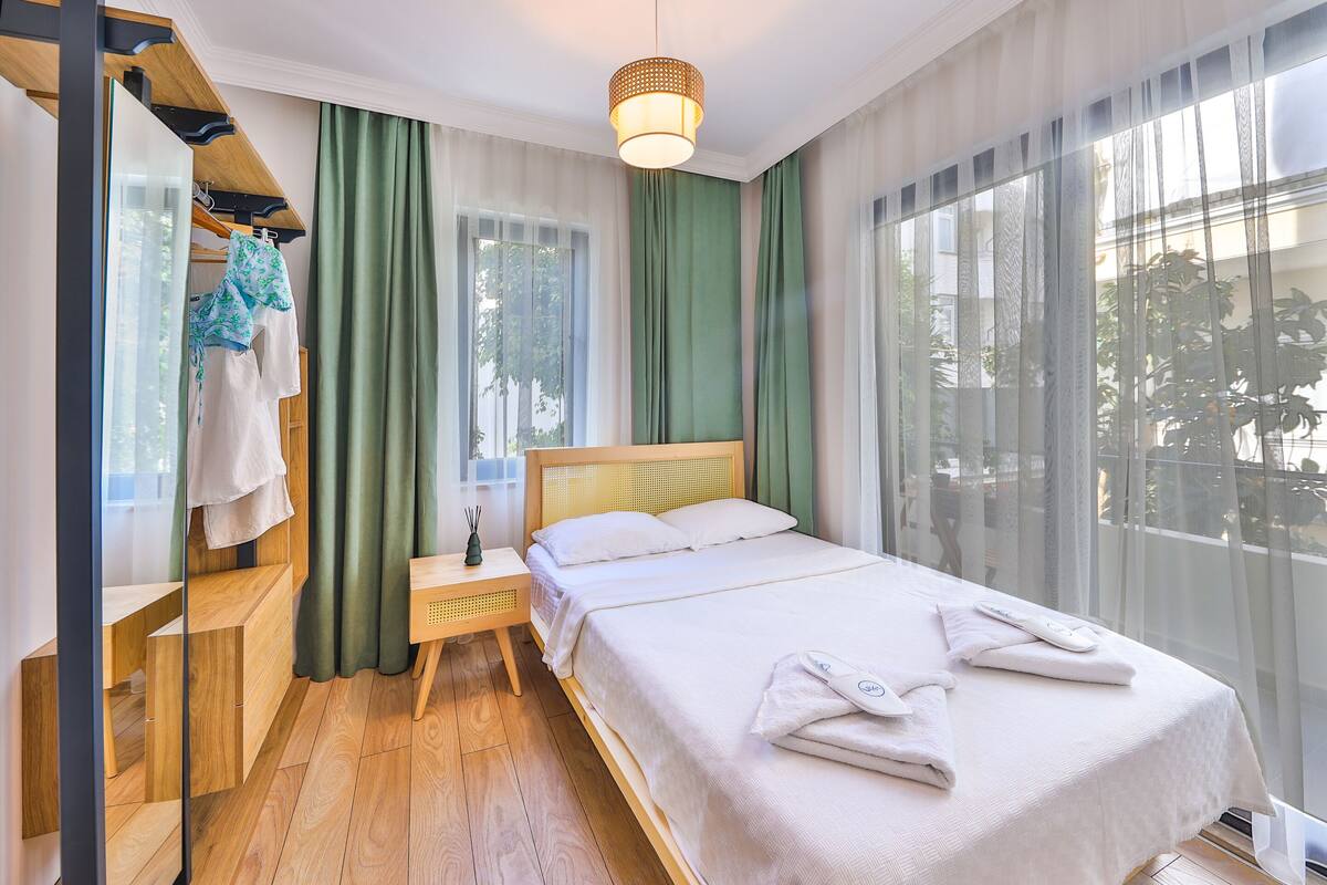 A hotel room with a wooden bed, green curtains, a wooden nightstand, and a hanging robe.