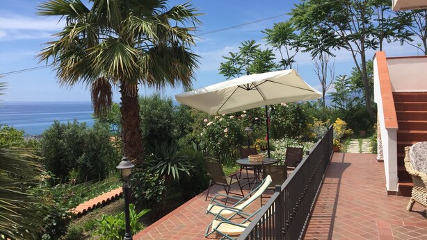 Een terras met een tafel, stoelen en een grote parasol, met uitzicht op de zee.