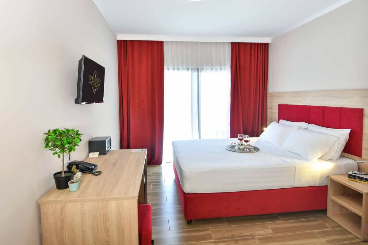 A hotel room with a bed, a desk, a telephone, a small plant, and a window with red curtains.
