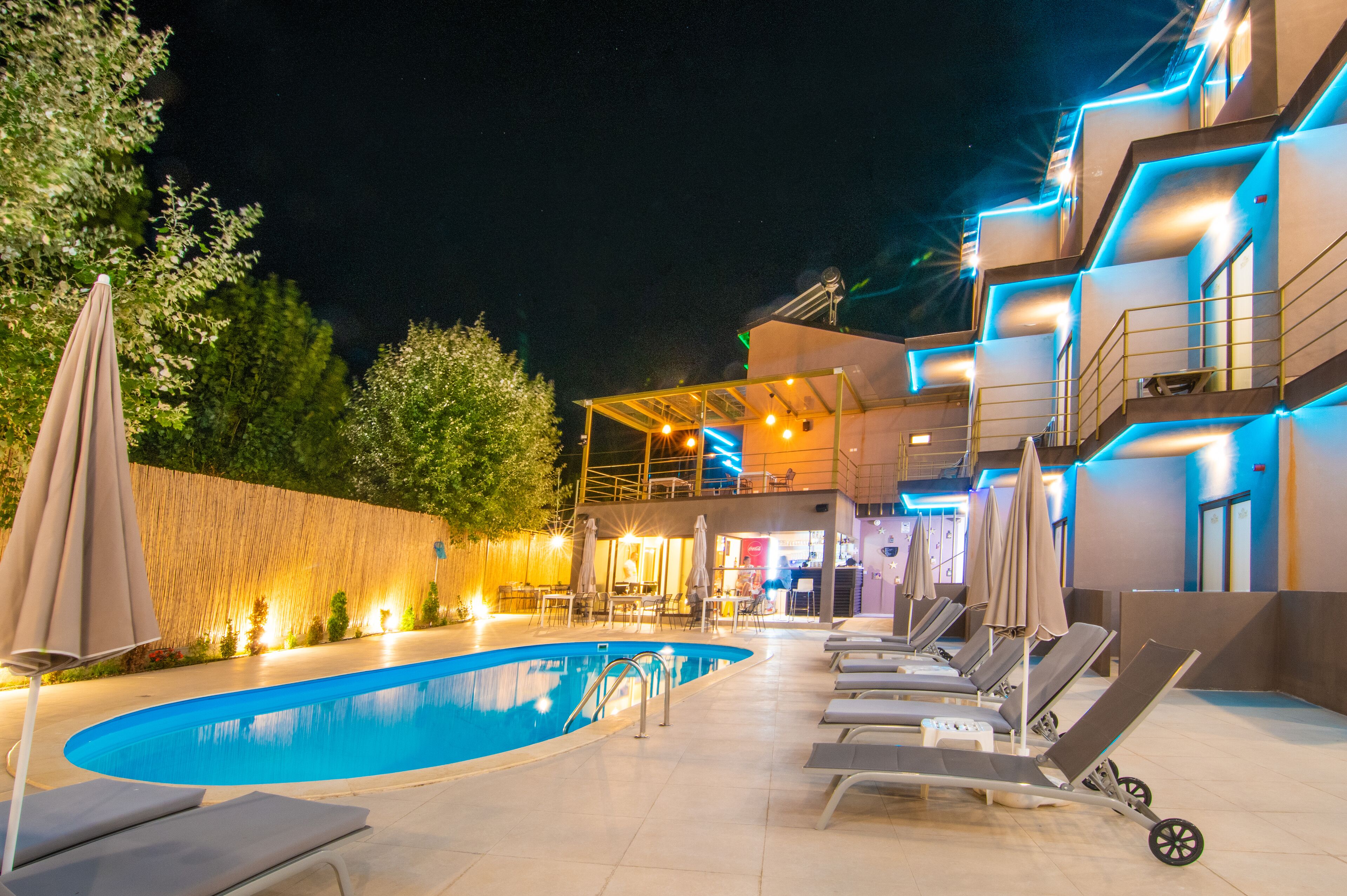 A swimming pool area with lounge chairs, umbrellas, and a building with illuminated balconies at night.