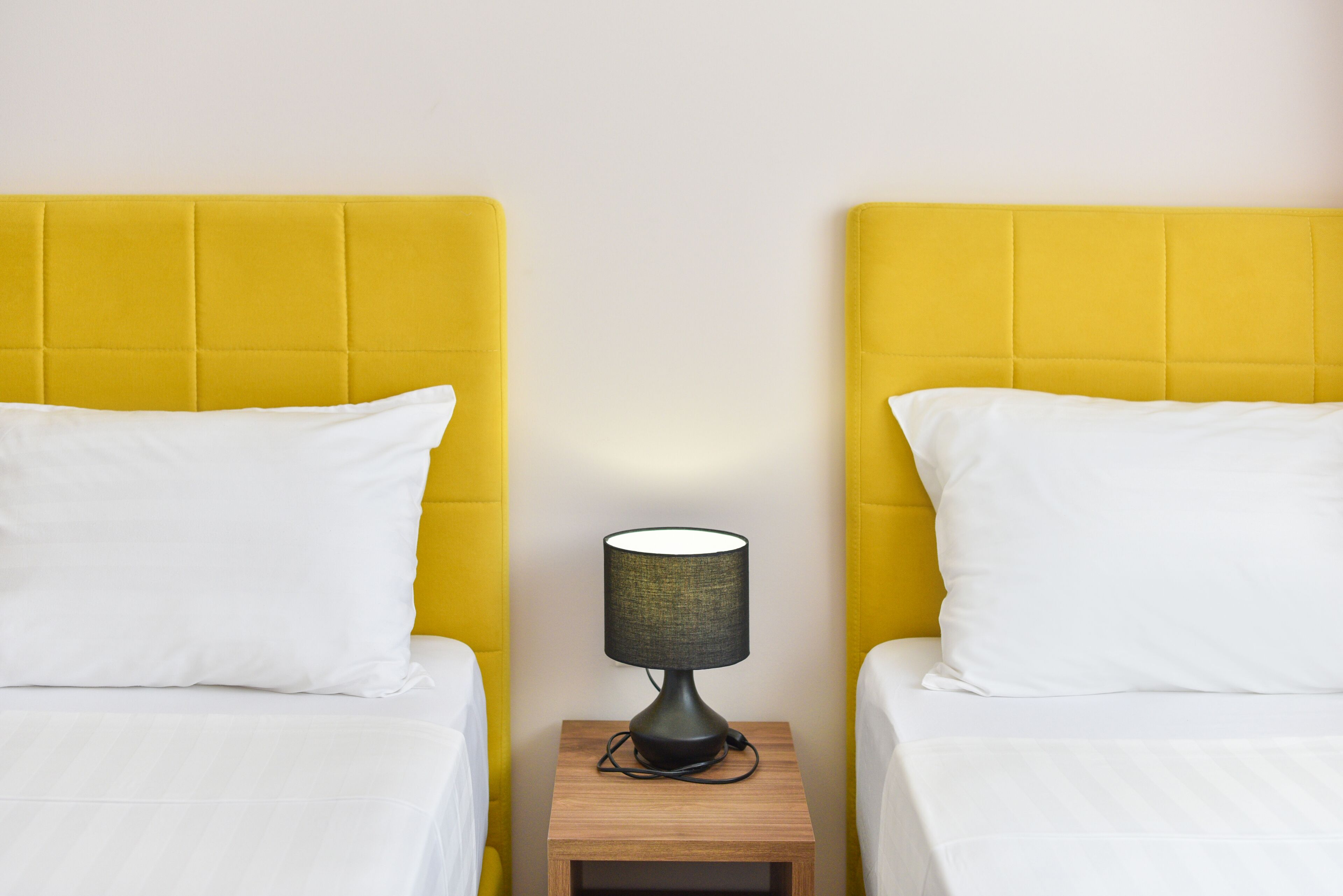 A room with two beds, each with a yellow headboard and white bedding. There is a small wooden nightstand with a lamp between the beds.