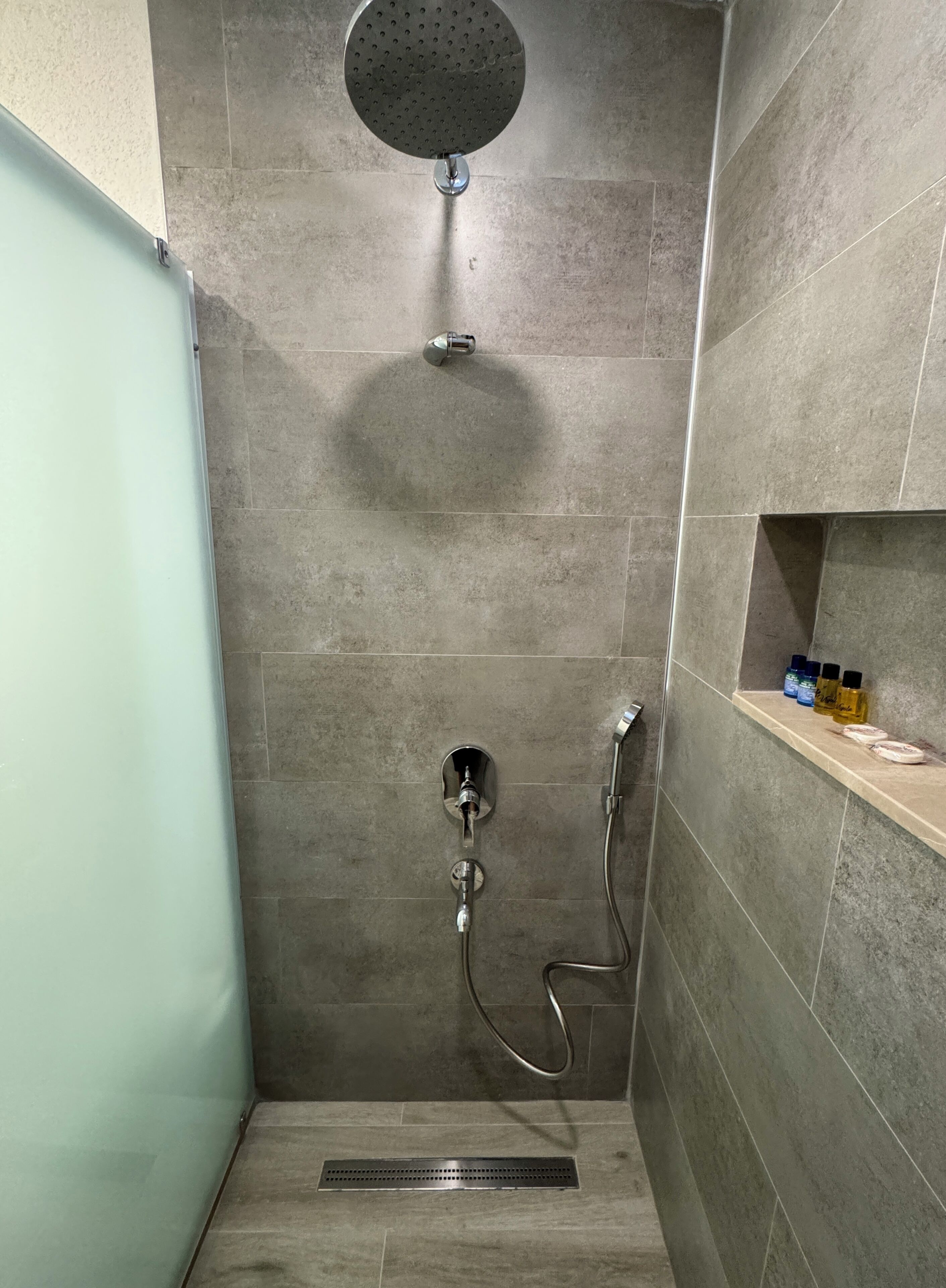 A shower with a handheld showerhead, a wall-mounted faucet, and a shelf with toiletries.