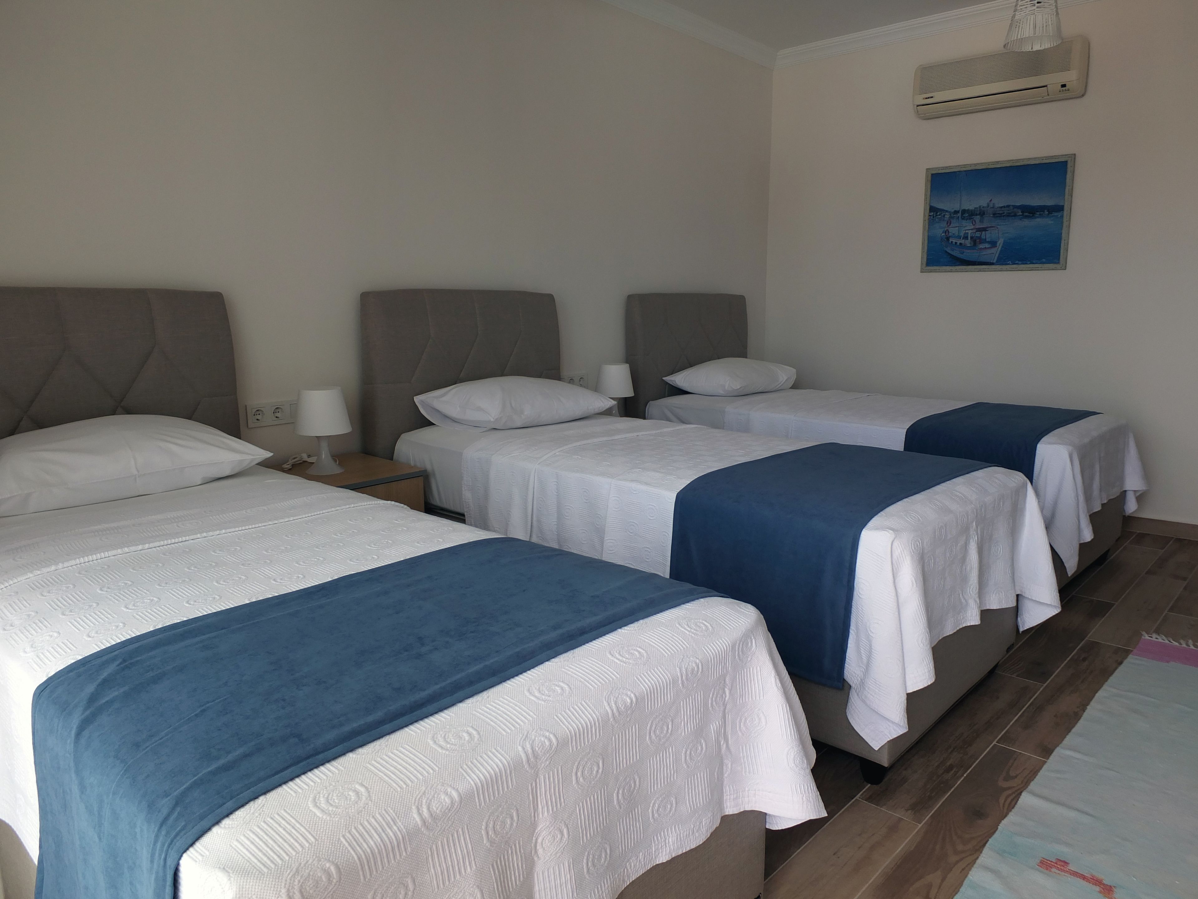Una habitación de hotel con tres camas, una mesita de noche con lámpara, aire acondicionado y un cuadro en la pared.