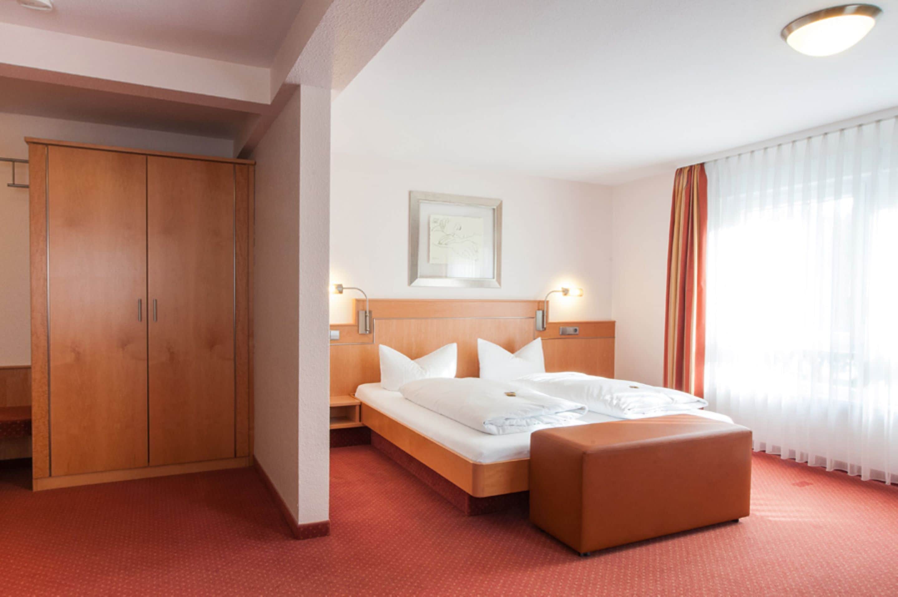 Ein Hotelzimmer mit einem Holzschrank, einem Bett mit weißer Bettwäsche, einem Nachttisch und einem Fenster mit durchsichtigen Vorhängen.