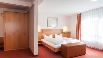 Ein Hotelzimmer mit einem Holzschrank, einem Bett mit weißer Bettwäsche, einem Nachttisch und einem Fenster mit durchsichtigen Vorhängen.