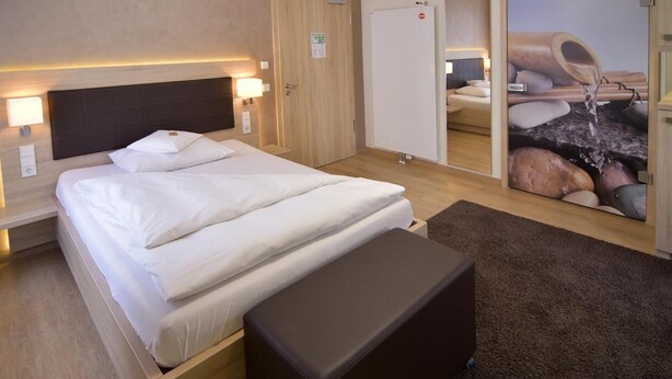 Quarto de hotel com uma cama grande, luminárias de cabeceira, um painel de madeira na cabeceira, uma mesinha de cabeceira pequena, um banco de cor escura, piso coberto por carpete, uma porta de madeira e uma obra de arte na parede retratando um recurso hídrico.