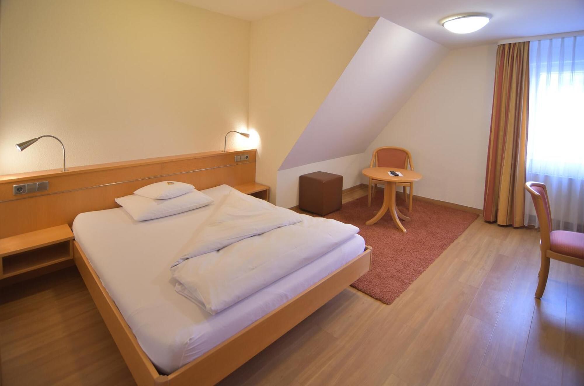 Et hotelværelse med seng, et natbord, en stol, et lille bord og et vindue med gardiner.