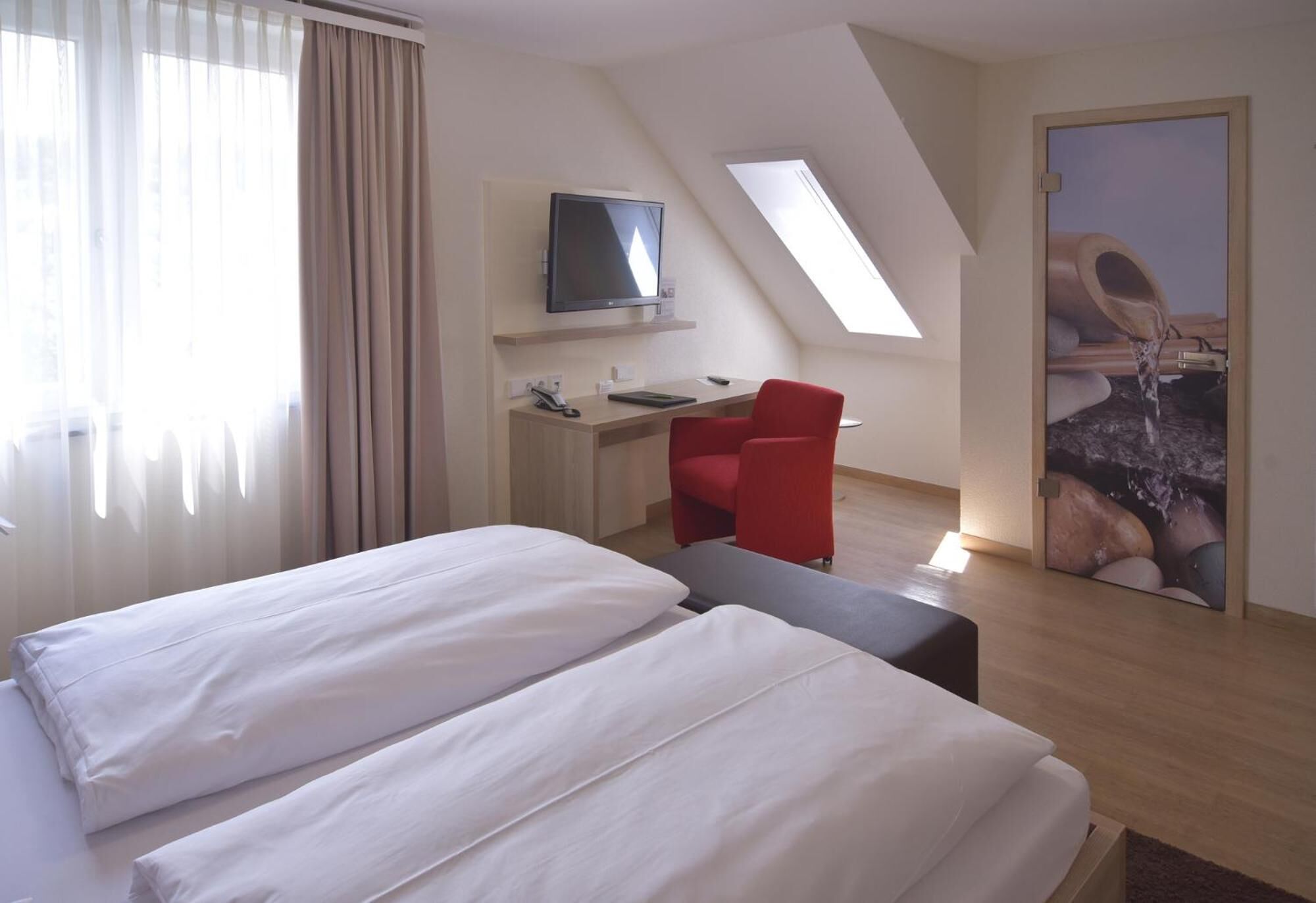 A modern hotel room with a bed, a desk with a computer, a red chair, and a television.