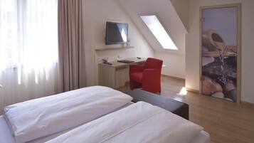 A modern hotel room with a bed, a desk with a computer, a red chair, and a television.