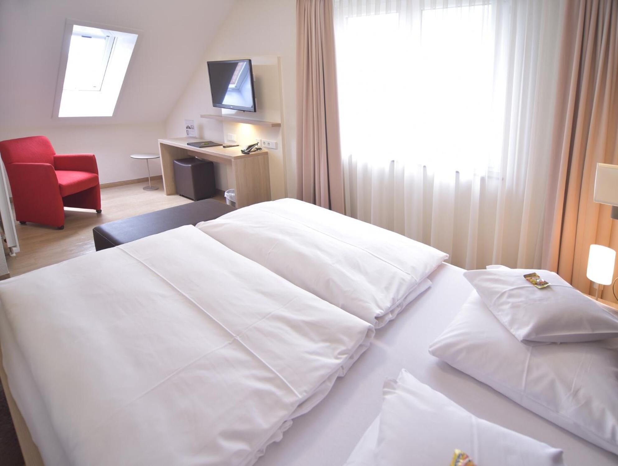 Une chambre d’hôtel avec deux lits, un bureau avec un ordinateur, une chaise rouge et un téléviseur fixé au mur.