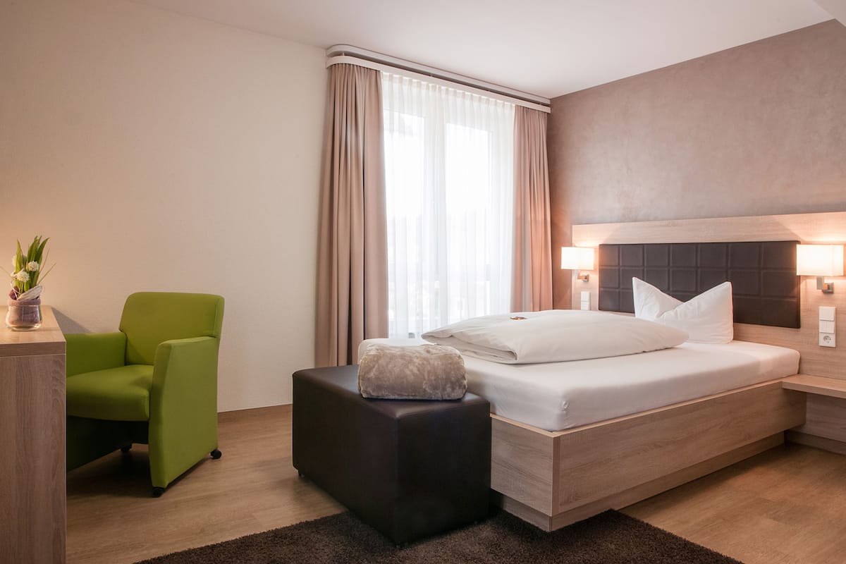 Una habitación de hotel moderna con una cama, una silla verde, un pequeño taburete, una mesita de noche y una lámpara.