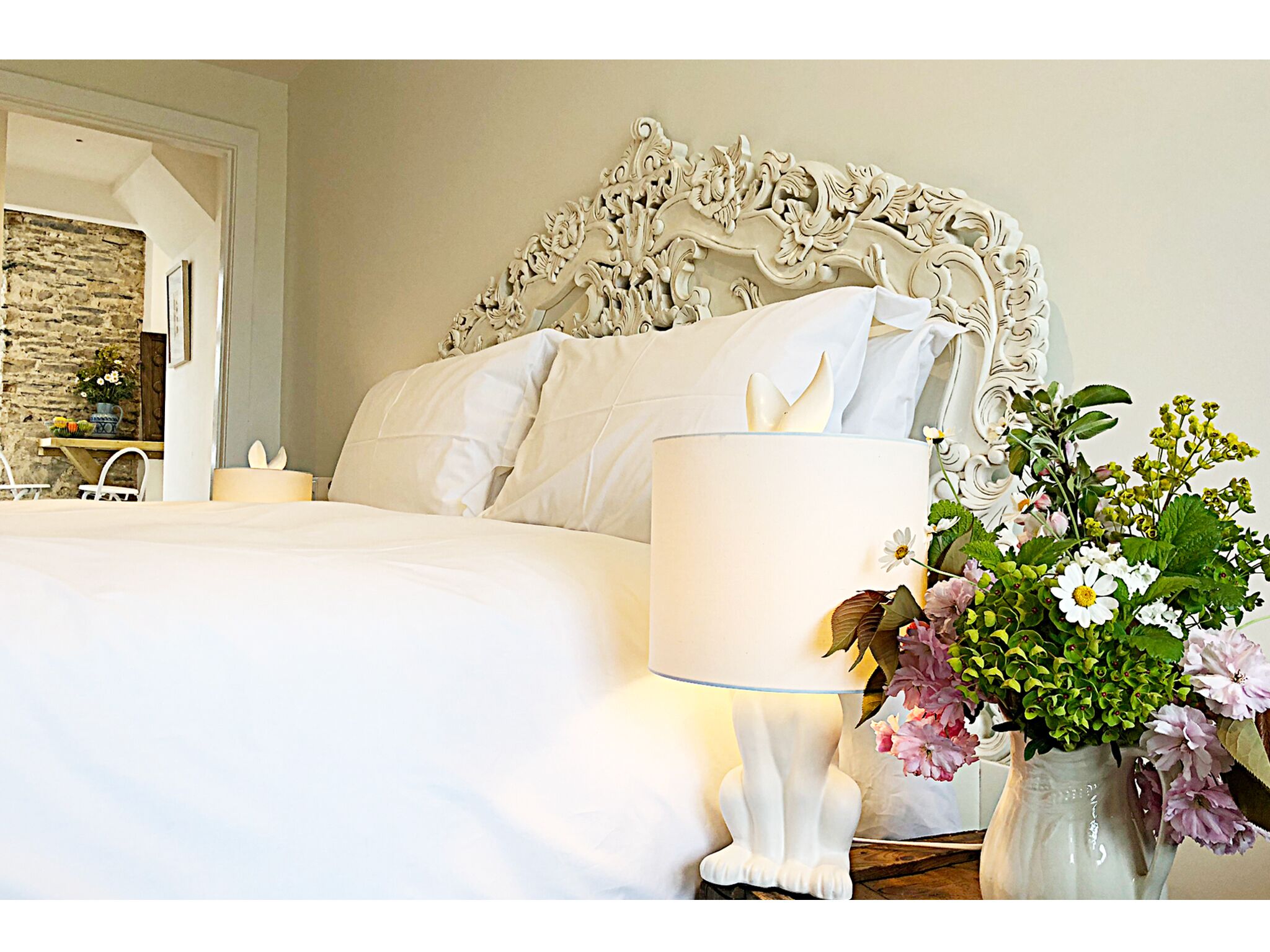 Una cama bien tendida con sábanas blancas, una lámpara blanca y un jarrón con flores.