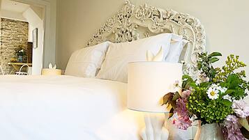 Una cama bien tendida con sábanas blancas, una lámpara blanca y un jarrón con flores.