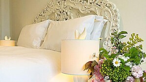 A neatly made bed with white linens, a white lamp, and a vase with flowers on a wooden nightstand.