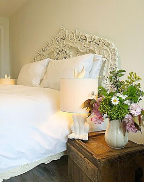 A neatly made bed with white linens, a white lamp, and a vase with flowers on a wooden nightstand.