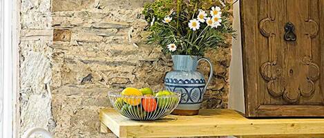A rustic stone wall, a wooden table with a bowl of fruit, a vase with flowers, and two white chairs.
