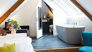 A modern attic bedroom with a sloped ceiling, a skylight, a large window, a bathtub, and a sofa with pillows.