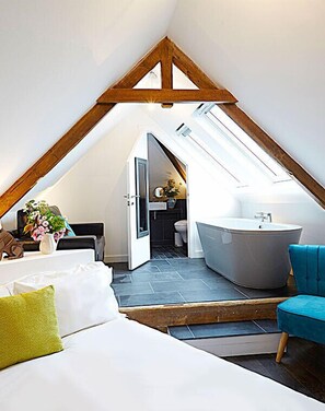 A modern attic bedroom with a sloped ceiling, a skylight, a large window, a bathtub, and a sofa with pillows.